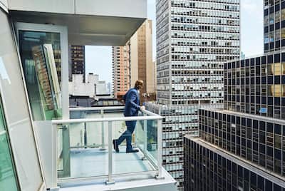 A man in a suit stands on a balcony looking out to a built up city view