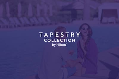 A laughing couple share a drink by the pool. The Tapestry Collection by Hilton logo is layered over the top of the image