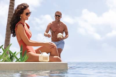Woman sits on edge of pool while man with drink walks towards her