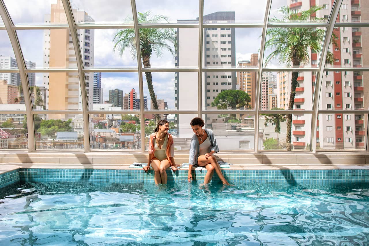 Two women sitting at the edge of the rooftop pool at Qoya Hotel Curitiba, Curio Collection by Hilton, with the city skyline in the background.