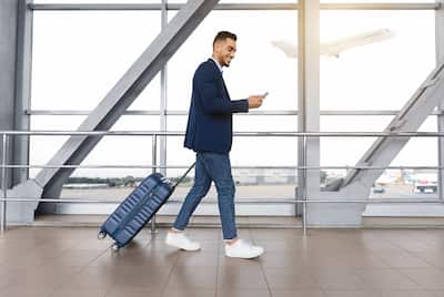 Young man with suitcase in airport looking at cell phone while smiling.